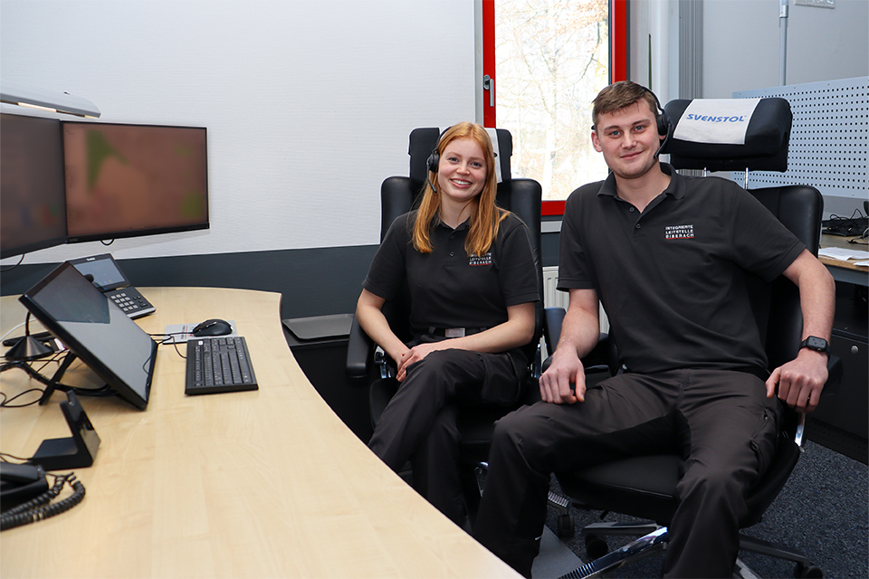 Ein junger Mann und eine junge Frau der Rettungsleitstelle mit Headsets lächeln in die Kamera.