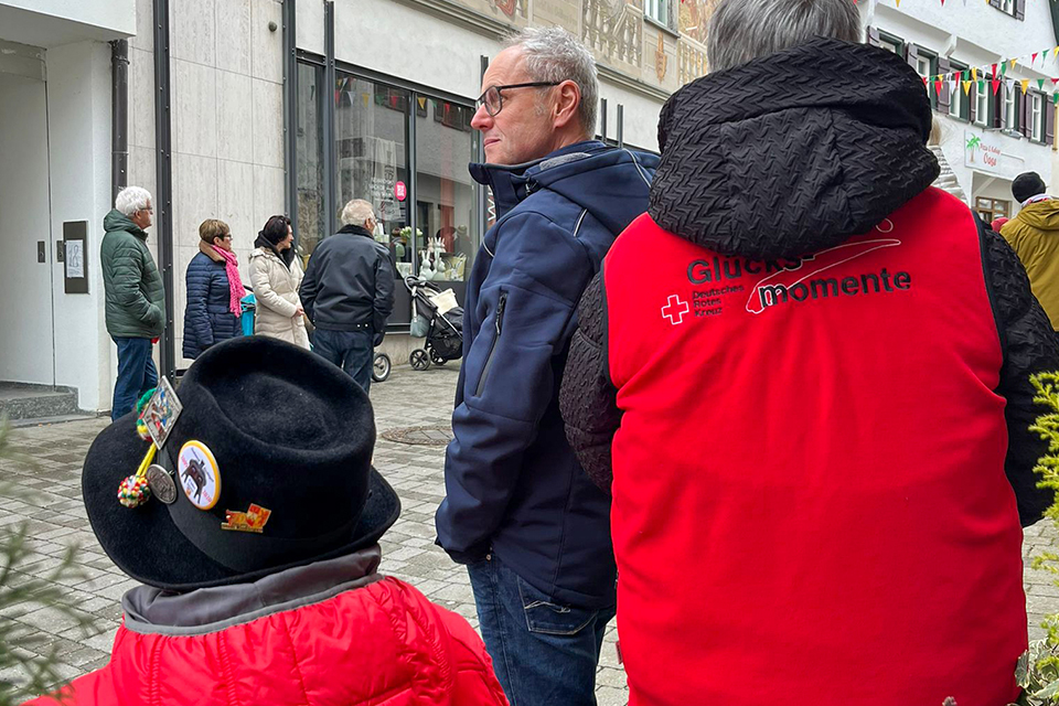 Mehrere Personen, unter anderem eine Rollstuhlfahrerin, sehen einem Faschingsumzug zu.